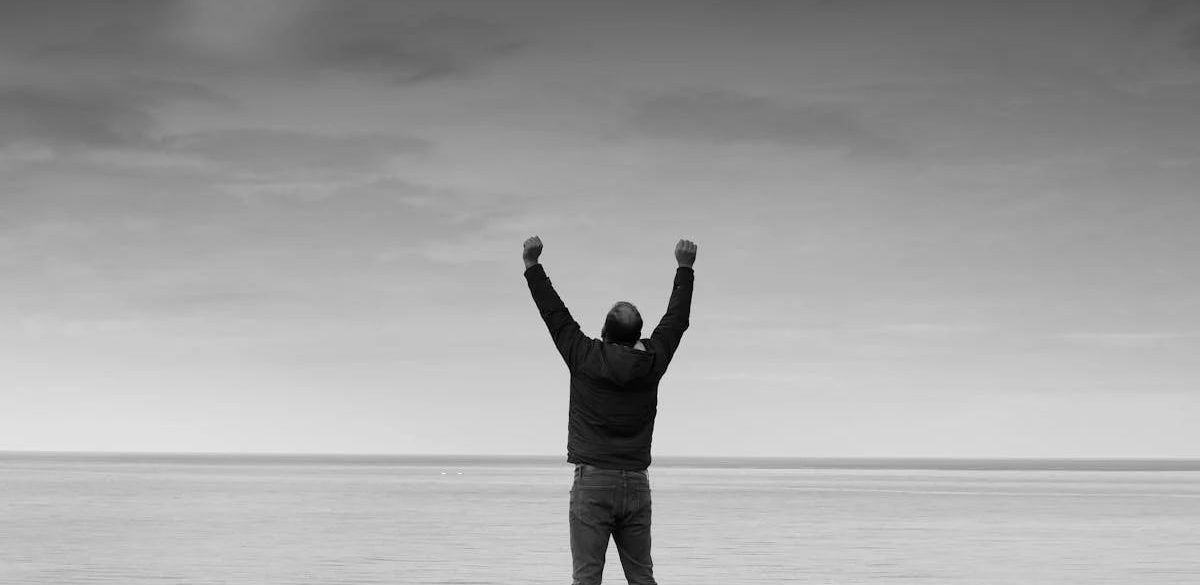 Black and white image of a person raising arms in celebration by the sea.