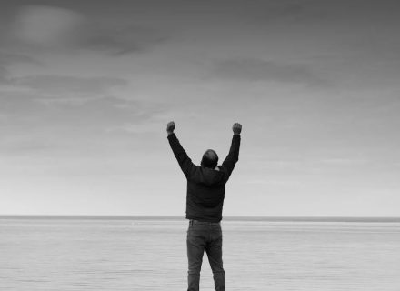 Black and white image of a person raising arms in celebration by the sea.