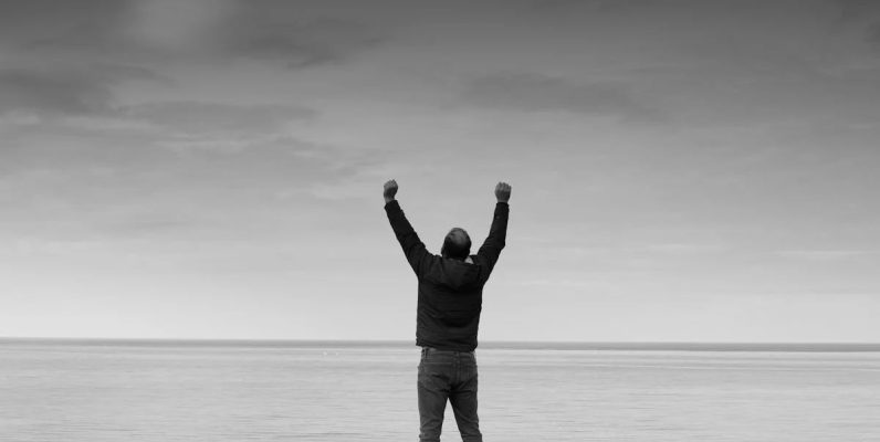 Black and white image of a person raising arms in celebration by the sea.